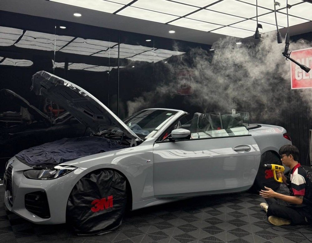 ช่างกำลังใช้เครื่องเป่าลมร้อนติดตั้งฟิล์มใสกันรอยกับรถยนต์ BMW สีเทาในห้องติดตั้งมาตรฐาน